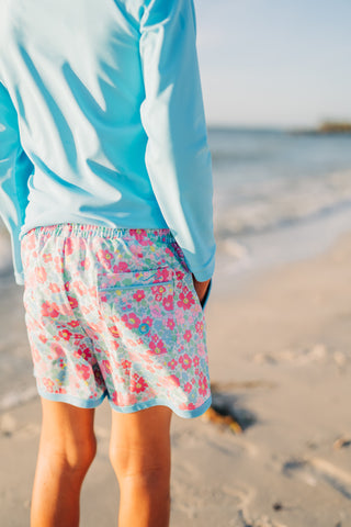 Person wearing a light blue jacket and floral shorts on a beach.