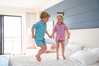 Two children in matching pajamas jumping on a bed in a bright bedroom.