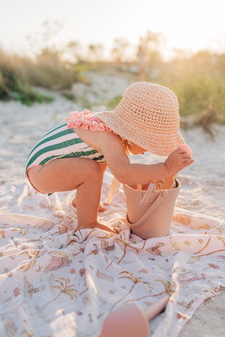Child on a beach wearing a striped swimsuit and sun hat, sitting on a patterned towel.