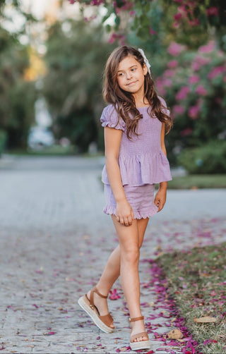 Young girl in a purple outfit standing in a garden with flowers and greenery.