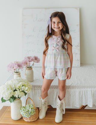 Young girl in a plaid outfit standing in a room with flowers and a painting on the wall.
