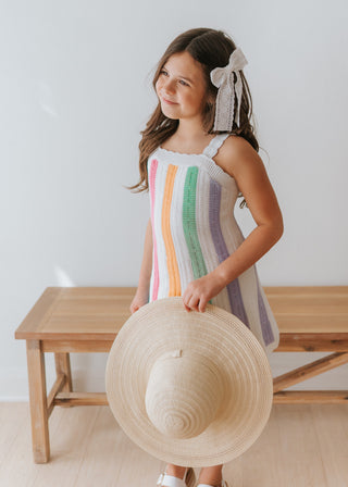 Young girl in a striped dress holding a straw hat, standing on a wooden floor with a white wall background.