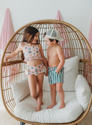 Two children in swimsuits standing on a wicker chair with a white cushion.