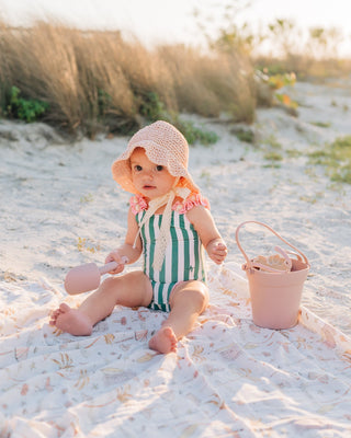 Baby in a green and white striped swimsuit and pink hat sitting on a blanket with a sand bucket in a sandy area.