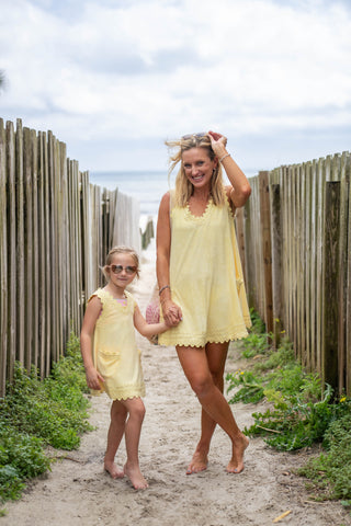 Two people in yellow dresses standing on a sandy path with wooden fences and plants in the background.