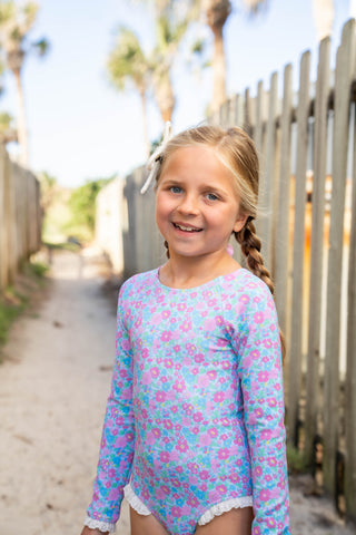 Young girl wearing a floral swimsuit standing on a beach path with palm trees in the background.