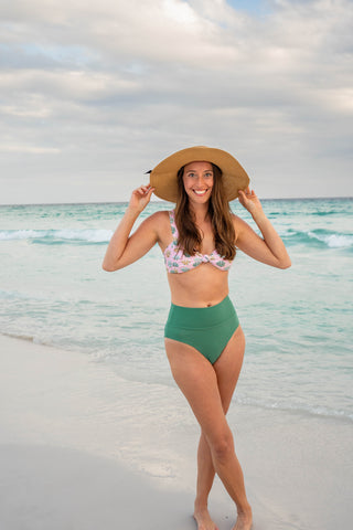 Woman in a green bikini and straw hat standing on a beach with ocean and sky in the background