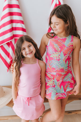 Two young girls wearing pink outfits standing next to each other with a striped towel in the background.