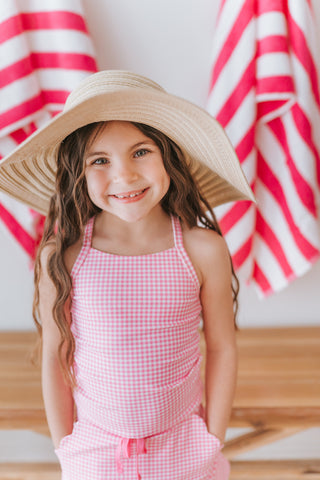 Young girl wearing a wide-brimmed hat and pink checkered outfit in front of red and white striped curtains.